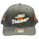 A dark gray snapback cap with 'JIG GO THUMP' embroidered on the front, resting on a wet dark wooden surface with a blurred background.