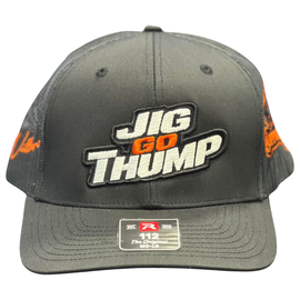 A dark gray snapback cap with 'JIG GO THUMP' embroidered on the front, resting on a wet dark wooden surface with a blurred background.